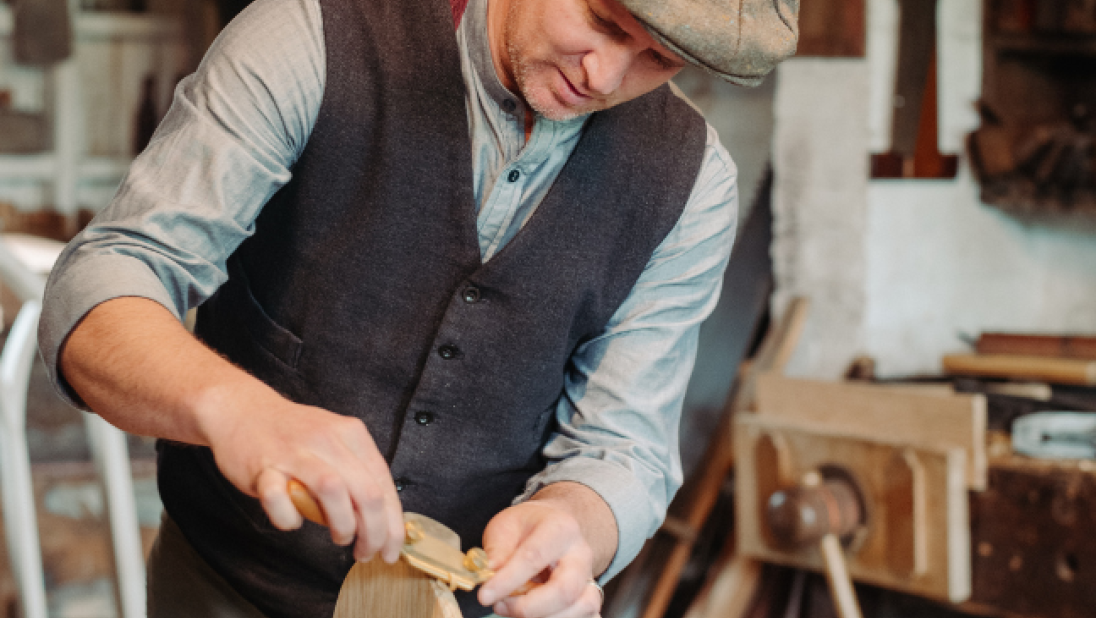 A heritage carpenter in 1900s dress - peaked cap, wool waistcoat, rolled up sleeves shirt - performing wood working skills.