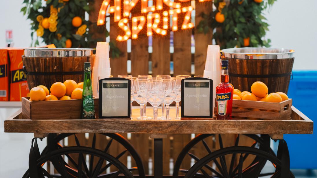 A cart with fruit and drink on it, with an orange lit up sign behind it saying Aperol Spritz. The sign has a foliage and oranges installation around it too.