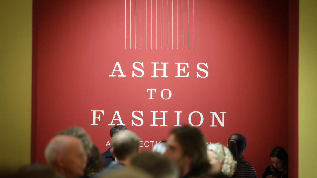 White text on a wall saying Ashes on one line, to in the middle line, then fashion underneath that on another line. There are vertical white lines going down to the wall as well as part of the design. The wall is a deep red colour, with heads of the crowd mingling in shot.