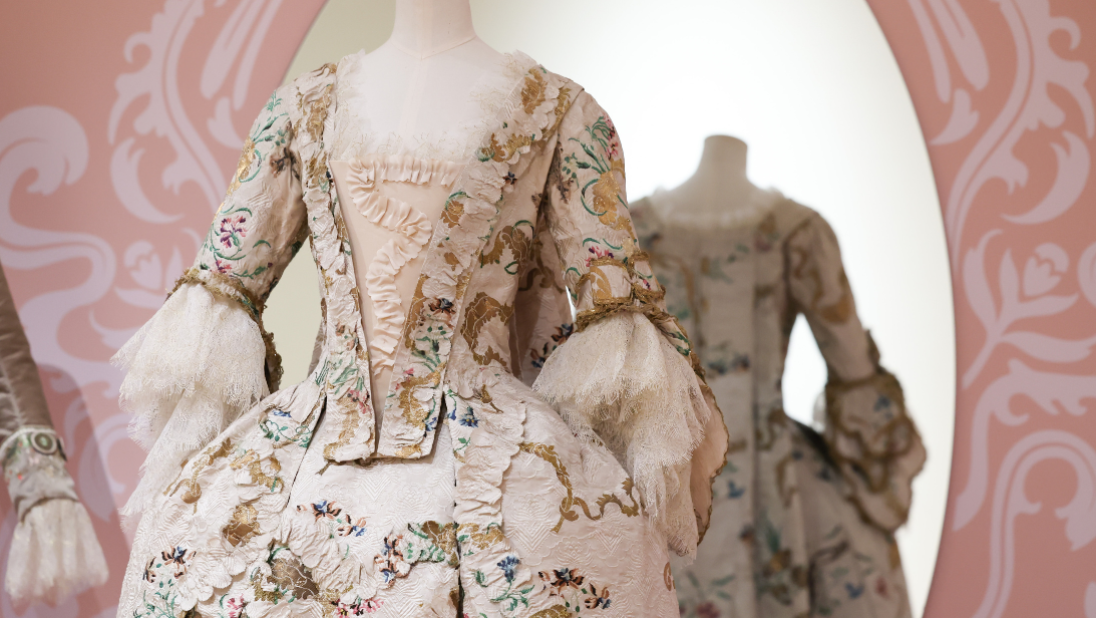 A cream and floral embroidered dress, with spirals of real gold thread, from the 1750s–1770s. It sits in front of a pink wallpaper with oval mirror, showing the back of the dress in the reflection.