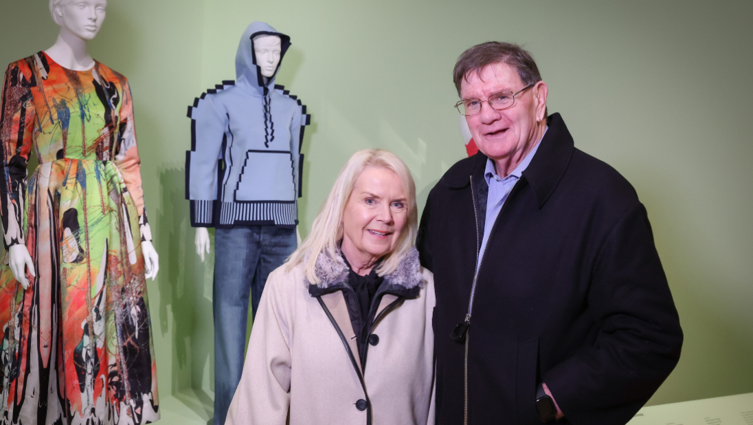 A couple standing in front of two outfits on mannequins, against a mint green wall.