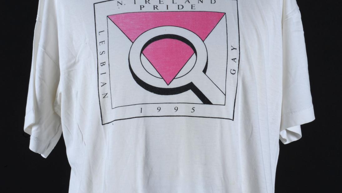 A white tshirt made for the fifth pride in Belfast. The logo has a pink inverted triangle and a magnifying glass.