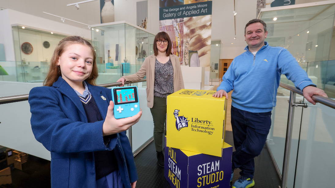 A girl dressed in blue school uniform, holding a games console and standing beside blue and yellow cubes that say Liberty IT and STEAM Studio. Representatives from the Ulster Museum and Liberty IT are pictured also, with the Applied Art Gallery in the museum as a backdrop.