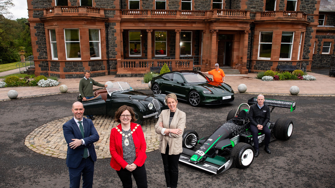 Three people, including a lady Mayor, standing in front of a manor house with a vintage car, new Porsche care and a race car parked outside with three other people standing at those. 