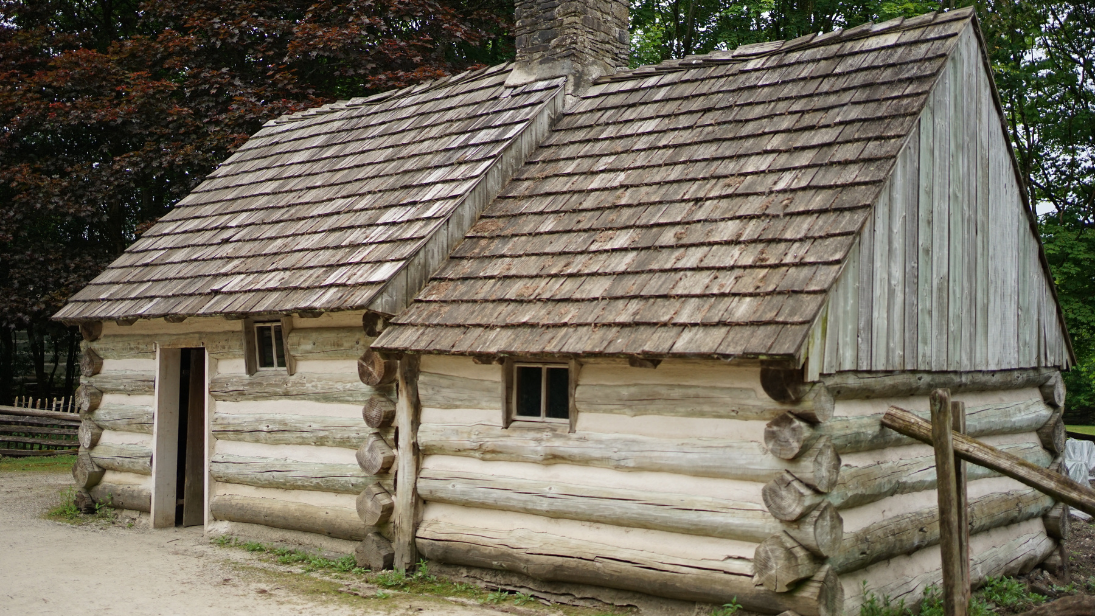 A log cabin, made of two rooms. Behind the cabin are large trees. To the left, the leaves are brownish red; to the right, they are green.