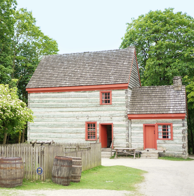 A log farmhouse with two storeys sits amidst some trees. The logs are varyingly blue and grey, while the trim is red.