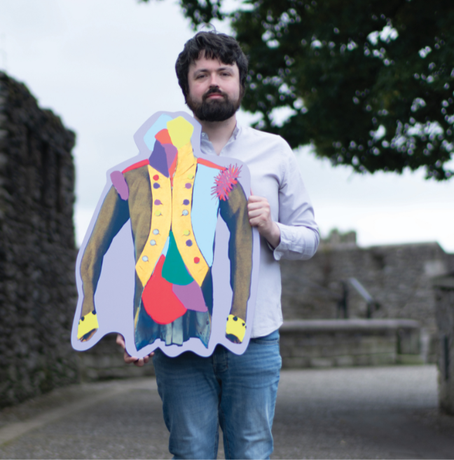 A male with dark hair and facial hair, wearing a grey long-sleeved polo and denim jeans, standing on Derry's Walls holding a cut-out of a colourful jacket museum object