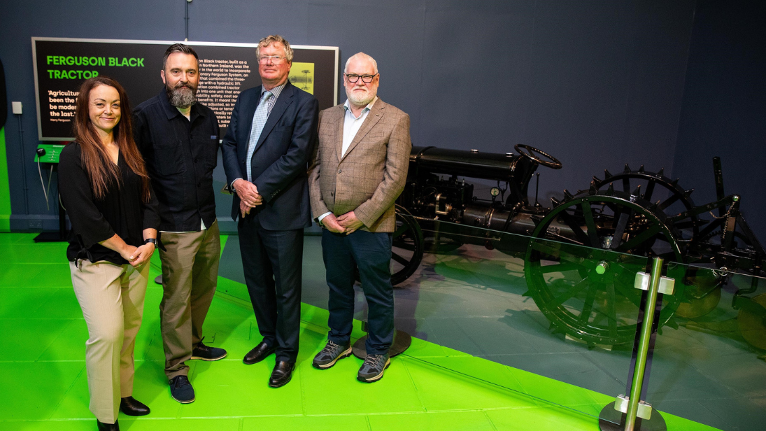 Four people standing in front of a black tractor which is displayed behind glass railings. The wall and part of the floor is painted a dark navy which joins another section of floor painted bright green.