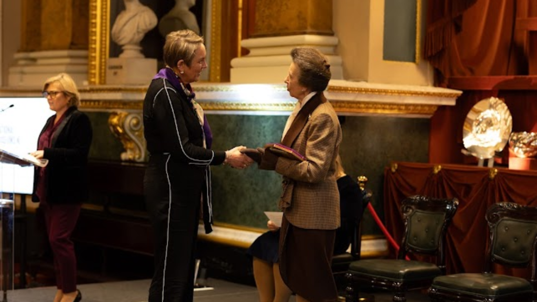 Chief Executive of National Museums NI, Kathryn Thomson, dressed in a black jumpsuit, shakes hands with Princess Anne as she receives an awards. Princess Anne wears a tan coloured blazer, neck scarf and gloves.