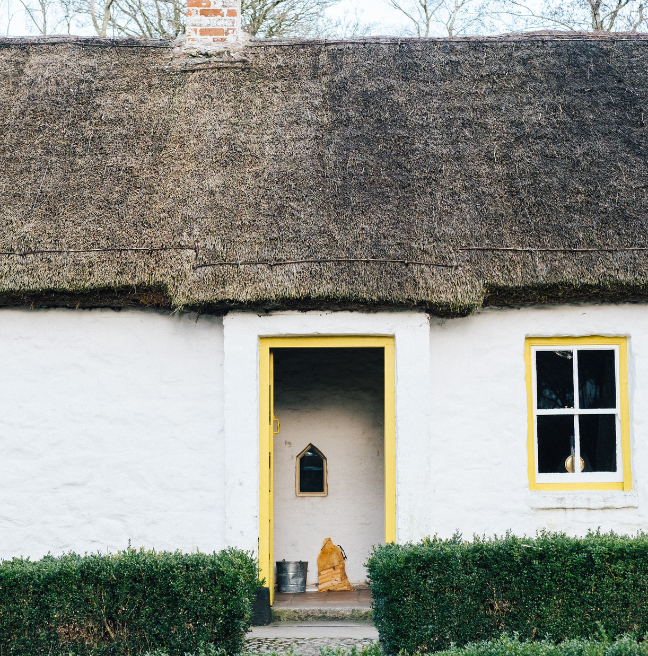 A white house with a yellow door stands open. The roof is thatched, and you can see the chimney and a blue-grey sky above. 