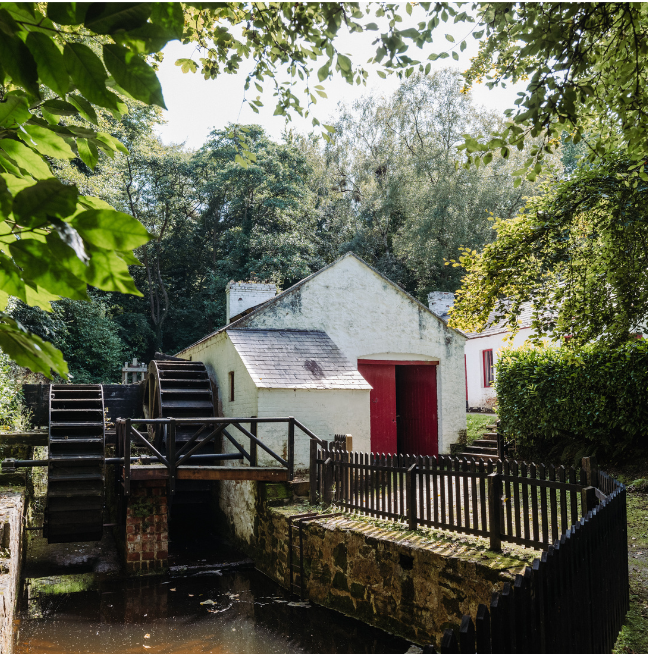 A white building sits nestled among trees; to its right is a water wheel. The door and window panes are bright red. 