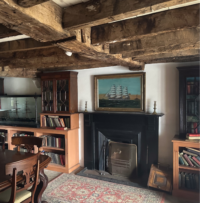 A view of a cosy room, with a table and chairs, a rug, books, and exposed wooden beams on the ceiling.