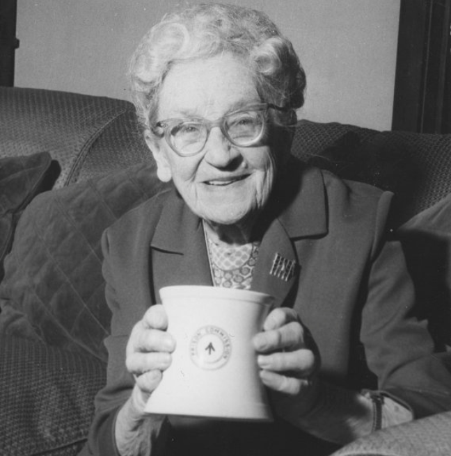 A smiling, older woman holds up a commemorative mug that celebrates the women's suffrage movement.