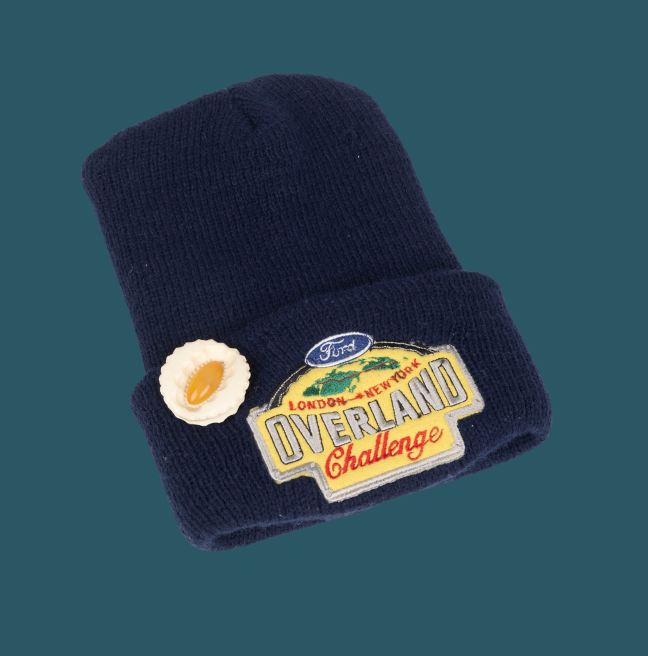 A blue beanie hat with a patch that reads 'Ford London - New York Overland Challenge'. There is a brooch shaped like an oyster attached.