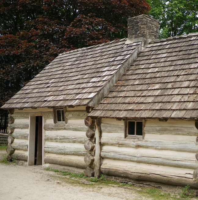 A log cabin, made of two rooms. Behind the cabin are large trees. To the left, the leaves are brownish red; to the right, they are green.