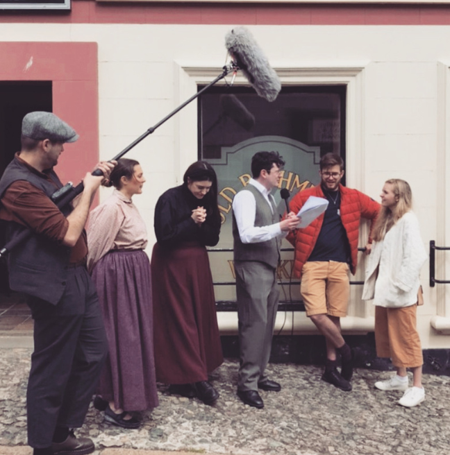 Group of men and women in period costume interviewing couple. Visable boom