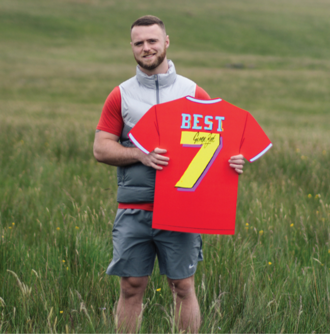 A young male with dark hair and beard standing in a grassy area of a mountain wearing grey shorts, grey gilet and a red t-shirt holding an artworked cut out of George Best's Man Utd shirt with Best written in blue and no. 7 in yellow.