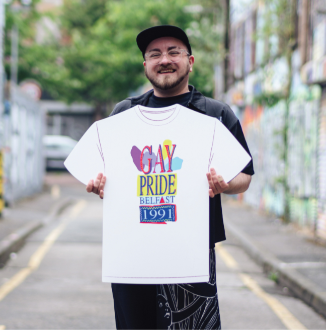 A person wearing glasses and wearing a baseball cap, wearing all black/denim. Holding an artwork cut-out of a Gay Pride t-shirt.