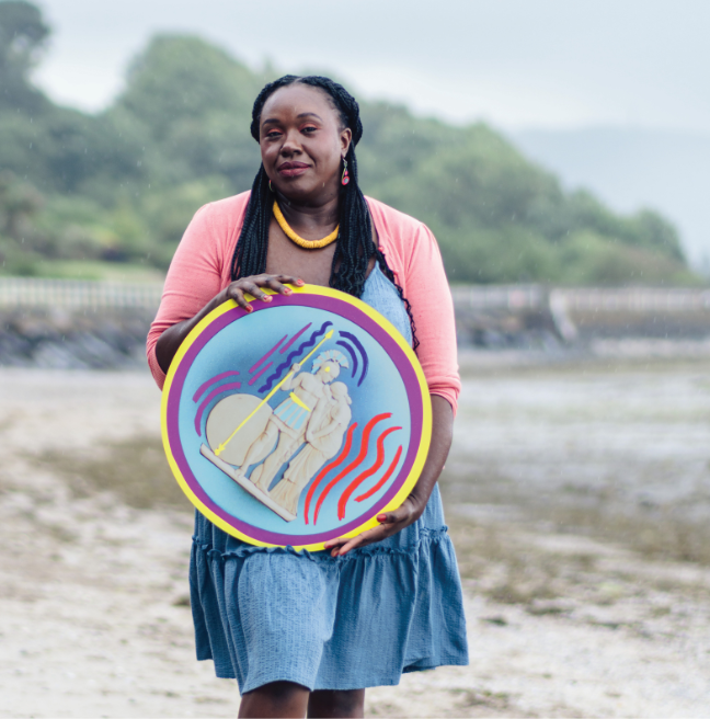 A black woman, with braided hair, standing on a beach wearing a pink cardigan over a blue denim dress and she has a yellow beaded necklace on too.  She is hold a round cut-out of an object from the Ulster Museum