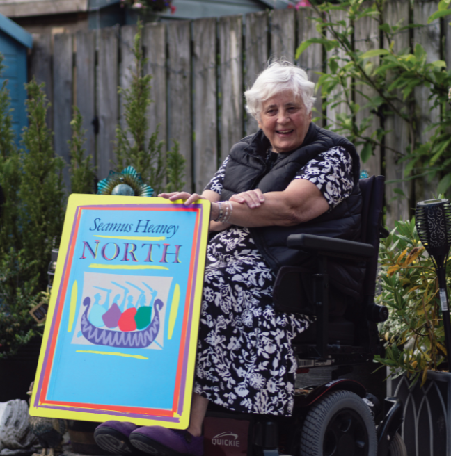 A person sitting in a wheelchair, in her garden, wearing a flowery dress and has white hair. She is holding a cut-out of a museum object that is blue in colour and says Seamus Heaney North on it