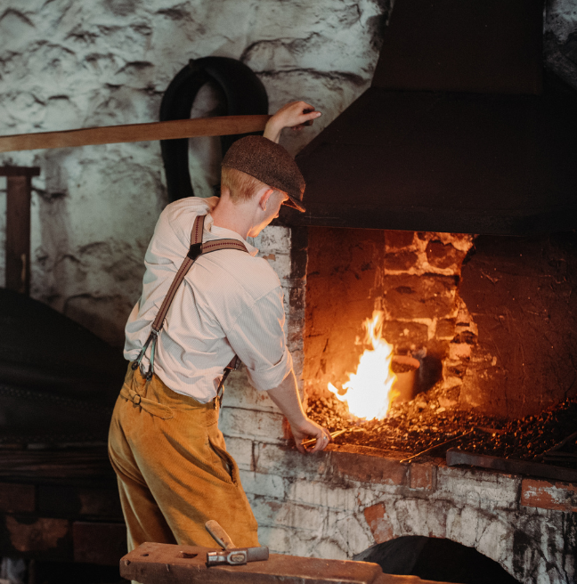 A blacksmith stands with his back to the camera, facing the fire of the forge.