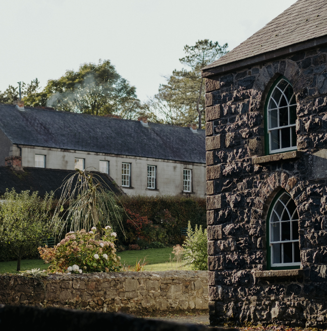 A peek at the Ulster Folk Museum, with a stone school on the left with arched windows, and a garden and a row of houses in the background peeking over a hedge.