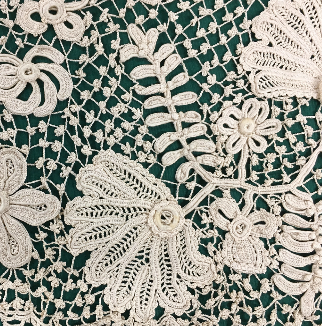 A close up vide of white lace on a green background. The lace has small knots in its grid, with beautiful floral detailing.