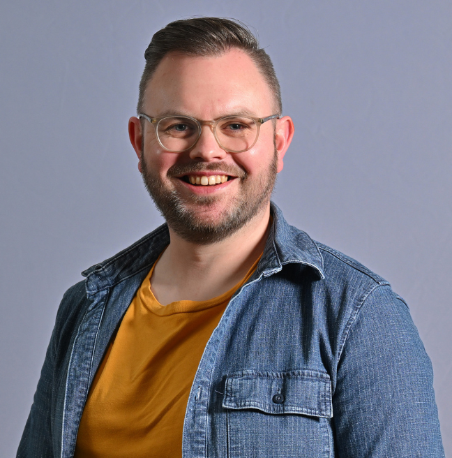 A man smiling at the camera with a plain grey photo backdrop behind him, wearing a denim shirt that's open with a mustard coloured t-shirt underneath. He wears glasses and has a short beard.