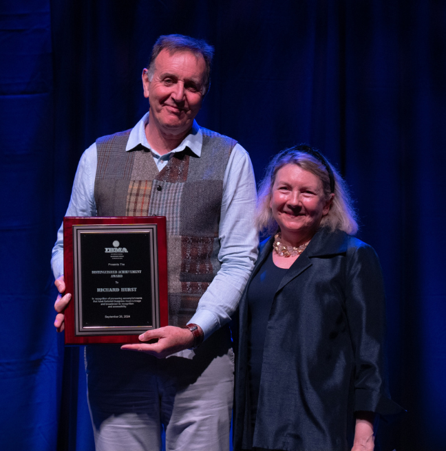 A man dressed in a shirt and waistcoat holding an award plaque beside a smaller in height woman, with shoulder length fair hair wearing a black outfit. There is a blue curtain as the backdrop.