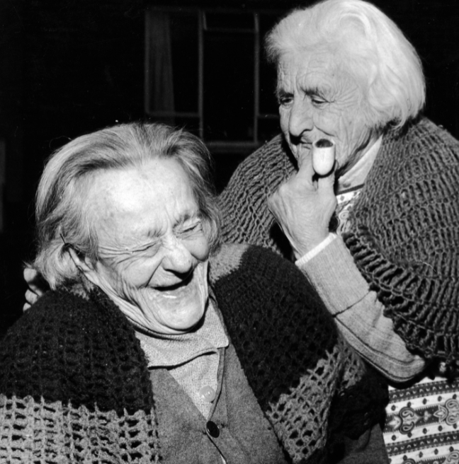 Two older women share a laugh together in a black and white photograph.