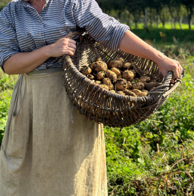 A woman dressed in linen clothes carries a woven basket filled with potatoes.