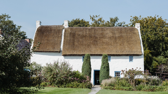 In a quiant garden, a large two-storey whitewashed and thatched cottage is flanked by two large hedges.