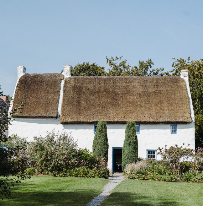 In a quiant garden, a large two-storey whitewashed and thatched cottage is flanked by two large hedges.
