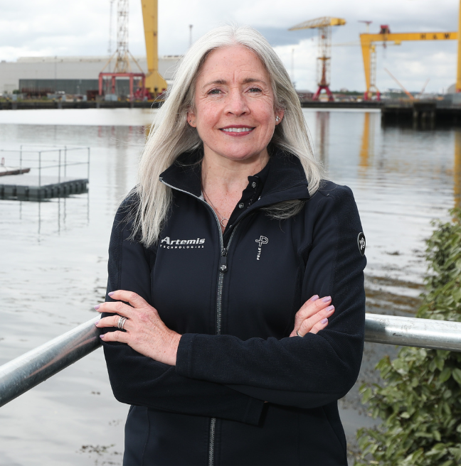 Professor Katrina Thompson standing with her arms crossed in Belfast harbour, Harland and Wolff cranes visible in the background