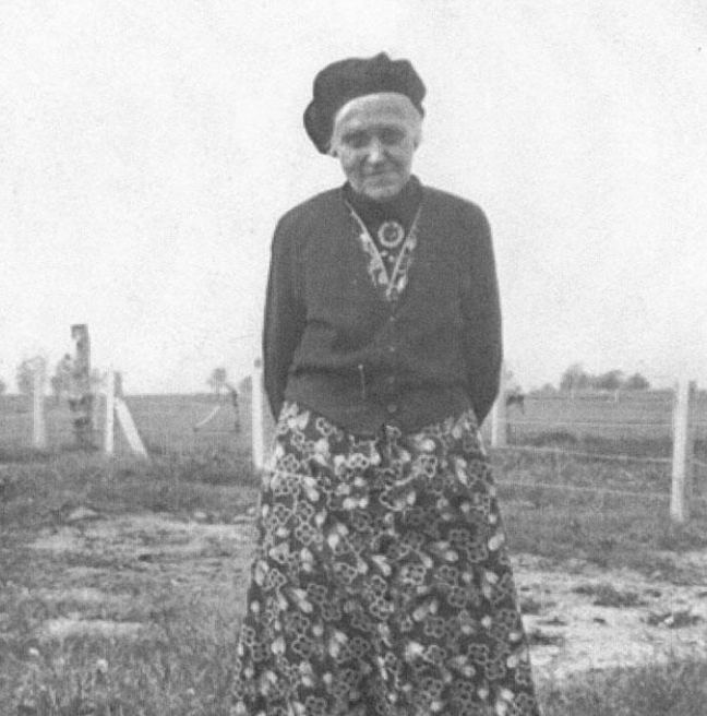 A smiling older woman looks at the camera in a black and white photograph. She is outdoors in a field environment, wearing a beret, patterned dress and cardigan.