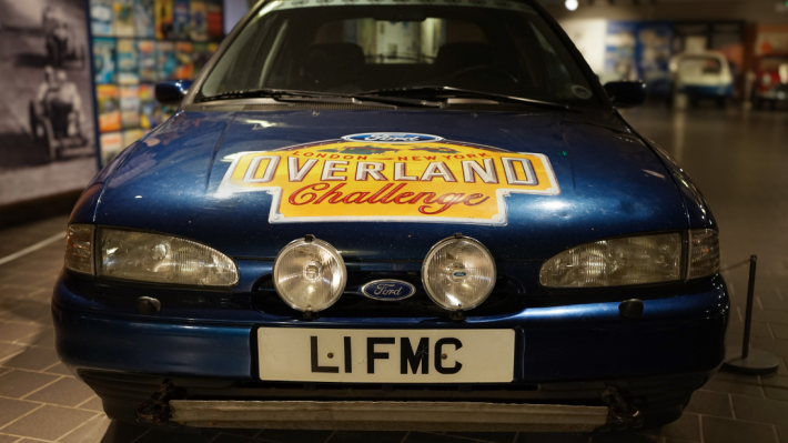 An image of the front of a car facing head on. It is a blue Ford, with the number plate L1 FMC and a large yellow decal reading 'Overland Challenge' on the hood.
