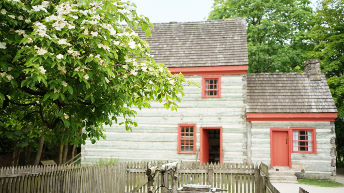 In the foreground of a photograph is a fenced garden and large tree. In the background is a large two storey farmhouse, with red trim and grey and blue logs.