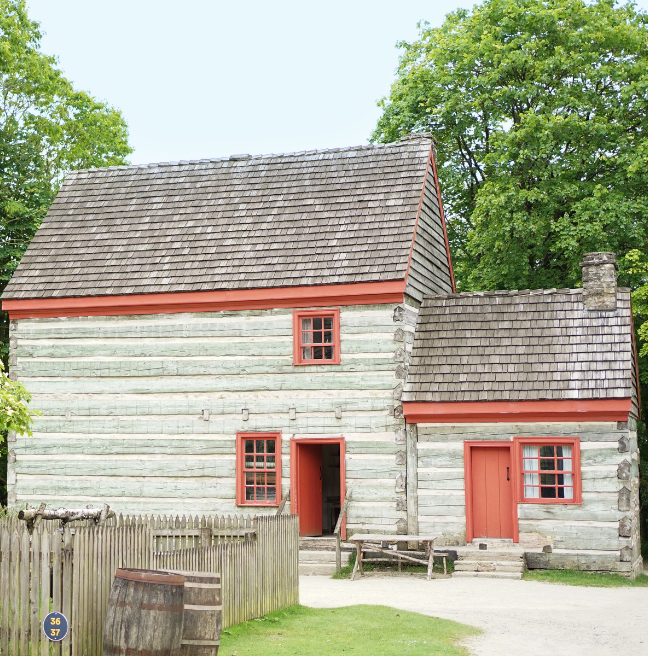 A large loghouse sits amongst trees. Its doors and window frames are a vivid red, while the logs have a grey and greenish hue.