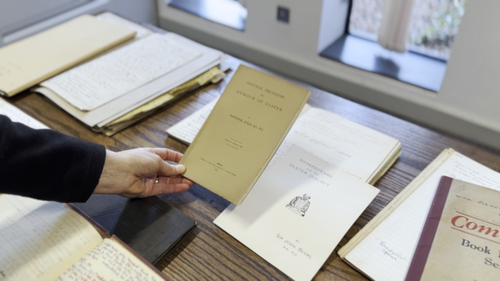 A woman's hand holds an old green pamphlet amidst other archival materials laid on a wooden table.