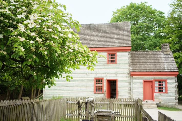 In the foreground of a photograph is a fenced garden and large tree. In the background is a large two storey farmhouse, with red trim and grey and blue logs.