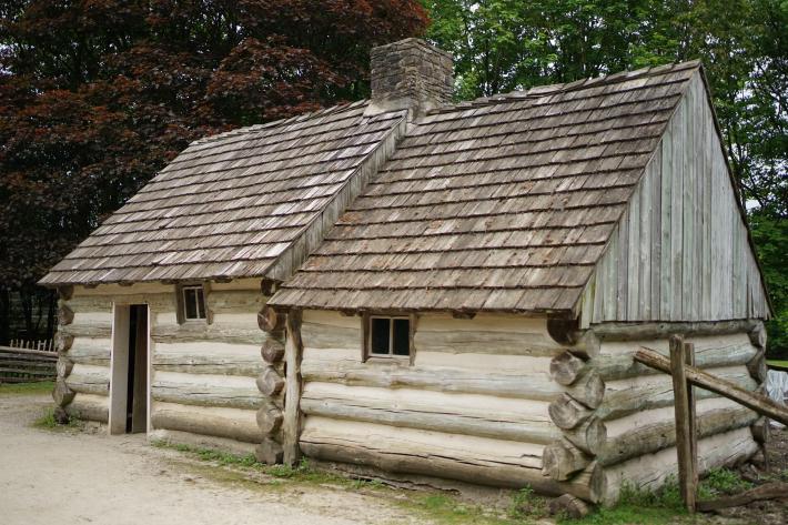 A log cabin, made of two rooms. Behind the cabin are large trees. To the left, the leaves are brownish red; to the right, they are green.