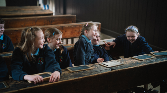 Six school children in their uniforms sitting at wooden benches and tables of an old style classroom in the Ulster American Folk Park. They're joking and smiling among themselves.