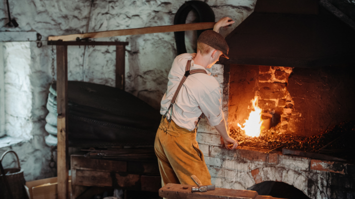 A blacksmith is at work in his forge. His back is to the camera, as he holds his tongs in the fire.