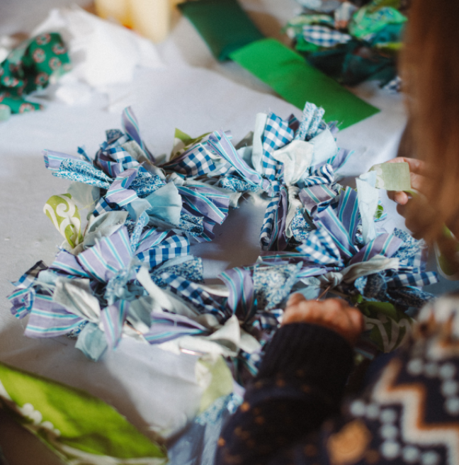 A woman just out of frame works on a blue rag wreath, tieing scraps of fabric to a circular frame. 