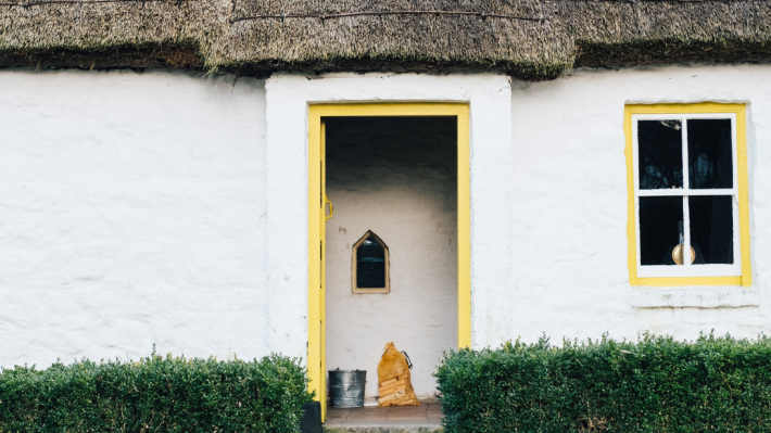 A whitewashed thatched cottage sits with its door open. The trim of the door is bright yellow. 
