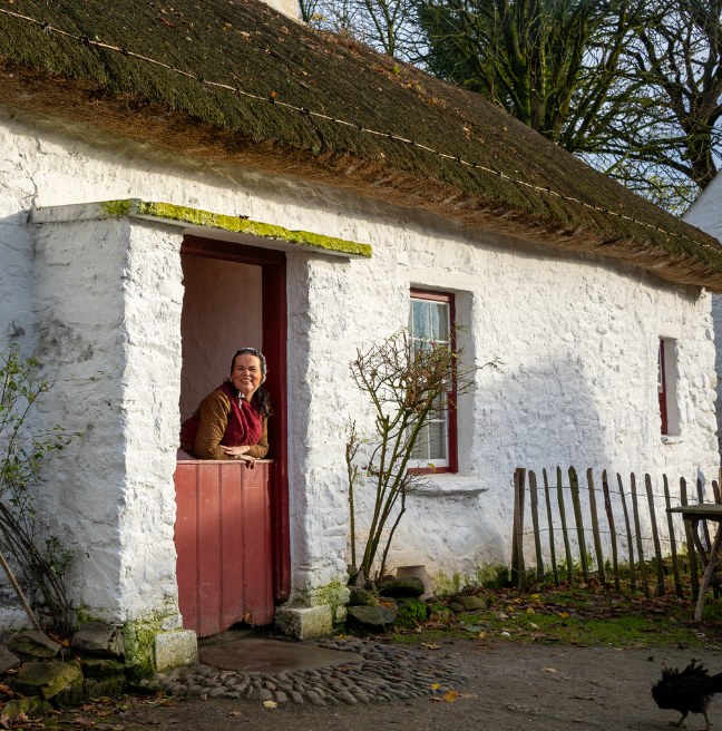 A woman stands in the doorway of a thatched cottage. She looks out at us, smiling.