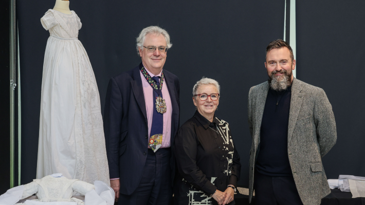 A white embroidered dress is on a mannequin in the background, to the left, and there people stand side by side, smartly dressed