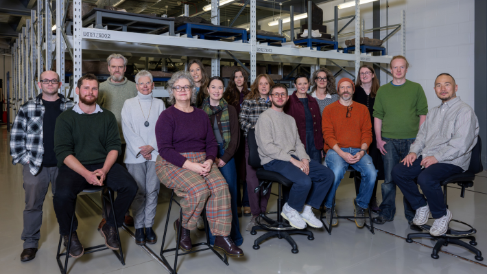 A group of 16 people, some seated, some standing, inside a museum store. The storage racks are in the background.