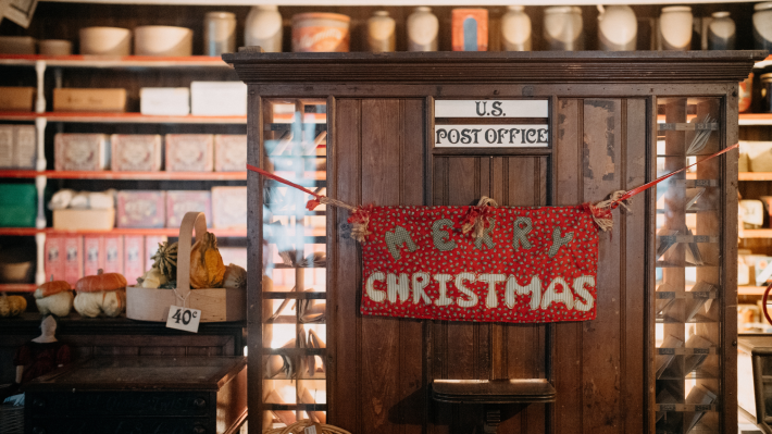 Inside an old times US Post Office. Lots of wood and parcels on shelves, a white sign says US Post Office in black. A red material banner with Merry Christmas letters stitched on it is hung from each corner of a wooden counter stall.