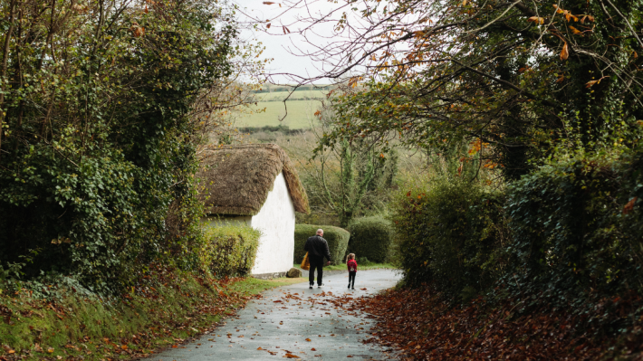A man and child walking with their backs to the camera on an autumn day past a white cottage 